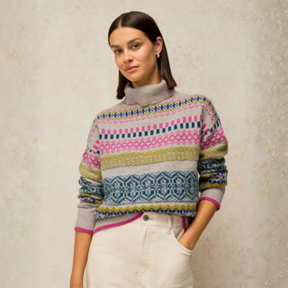 A woman wearing a multicolor Fair Isle patterned turtleneck sweater with pink, blue, yellow, and gray tones, paired with cream-colored pants, standing against a textured beige wall.