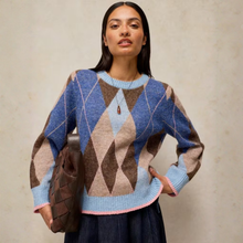 Load image into Gallery viewer, A woman models a blue, brown, and beige argyle knit sweater with light blue ribbed trim, holding a brown woven handbag against a textured beige wall.
