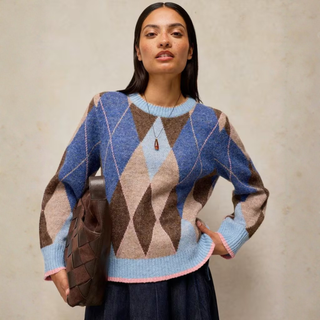 A woman models a blue, brown, and beige argyle knit sweater with light blue ribbed trim, holding a brown woven handbag against a textured beige wall.