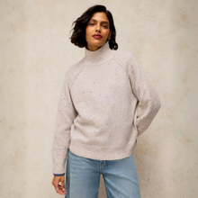 Load image into Gallery viewer, A woman wearing a light grey turtleneck sweater with subtle multicolor flecks and light blue jeans, standing against a textured beige wall.
