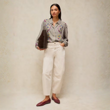 Load image into Gallery viewer, A woman stands against a textured beige studio backdrop wearing a striped button-up shirt with colorful floral embroidery, off-white cropped trousers, and burgundy loafers, while holding a dark brown clutch.
