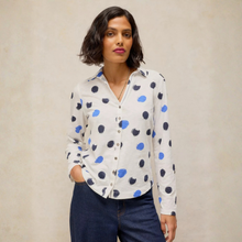 Load image into Gallery viewer, A woman modeling a white button-up blouse with blue and navy polka dots, paired with dark blue jeans, against a textured beige background.
