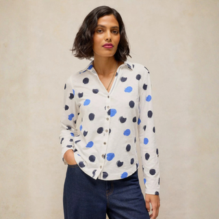 A woman modeling a white button-up blouse with blue and navy polka dots, paired with dark blue jeans, against a textured beige background.