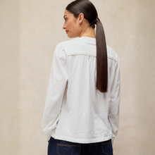 Load image into Gallery viewer, A woman with long dark brown hair in a ponytail, viewed from the back, wearing a white long-sleeve blouse with delicate lace trim at the shoulders and dark blue jeans, set against a neutral beige background.

