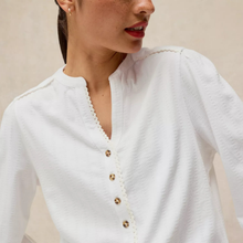 Load image into Gallery viewer, A close-up of a woman wearing a white textured blouse with a scalloped V-neckline and gold-tone buttons.
