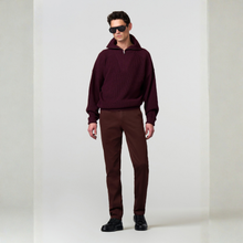 Load image into Gallery viewer, A man modeling a burgundy ribbed knit half-zip hoodie and matching burgundy trousers, styled with black sunglasses and black shoes, against a light gray studio background.
