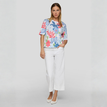 Load image into Gallery viewer, Woman wearing a colorful floral blouse and white pants on a gray background
