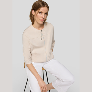 Woman wearing a beige jacket and white pants sitting on a stool against a plain background