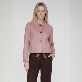 A woman modeling a dusty pink cropped suede jacket with three dark buttons and a pointed collar, paired with dark brown trousers, against a light gray textured studio background.
