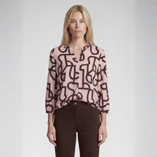 A woman modeling a light pink long-sleeve blouse with a dark brown abstract line pattern, paired with dark brown pants, against a light gray studio background.