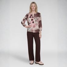 Load image into Gallery viewer, A woman modeling a long-sleeve abstract print blouse in pink, brown, and beige tones, paired with dark brown drawstring pants and light pink loafers, against a light gray studio background.
