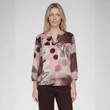 Load image into Gallery viewer, A woman modeling a long-sleeve blouse with an abstract print in shades of pink, brown, beige, and black, paired with dark brown trousers, against a light gray studio background.
