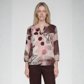 A woman modeling a long-sleeve blouse with an abstract print in shades of pink, brown, beige, and black, paired with dark brown trousers, against a light gray studio background.