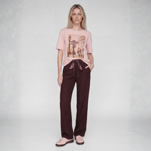 Load image into Gallery viewer, A woman stands against a light gray studio background wearing a light pink graphic T-shirt, dark brown drawstring pants, and matching pink slip-on shoes.
