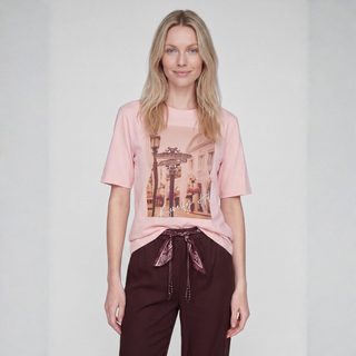 A woman modeling a light pink short-sleeve graphic t-shirt with a vintage street scene print, paired with dark brown drawstring pants, against a light gray studio background.
