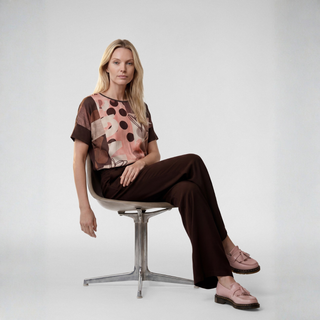 A woman sits on a modern swivel chair against a light gray studio background, wearing a short-sleeve abstract print top in brown, pink, and beige tones, dark brown pants, and dusty pink loafers.