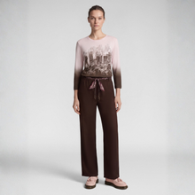 Load image into Gallery viewer, A model stands against a light gray studio background wearing a long-sleeve top with a pink-to-brown cityscape gradient and print, paired with dark brown wide-leg pants featuring a pink drawstring, and light pink flat shoes.
