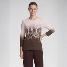 Load image into Gallery viewer, A woman modeling a three-quarter sleeve top with a gradient design that fades from light pink at the shoulders to dark brown at the hem and cuffs, featuring a black and white cityscape graphic across the chest. She is wearing dark brown pants against a light gray studio background.
