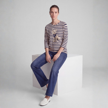 Load image into Gallery viewer, A woman sits on a white cube in a studio setting, wearing a navy and cream horizontal striped long-sleeve top with a dog graphic, blue jeans, and white loafers.
