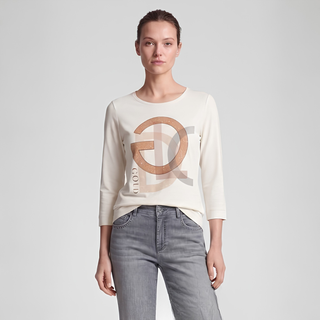 A woman modeling a cream-colored long-sleeve knit top with a large metallic gold 'GOLD' graphic on the front, paired with gray jeans, against a light gray studio background.