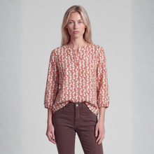 Load image into Gallery viewer, A woman modeling a loose-fitting, three-quarter sleeve blouse with a coral and beige geometric pattern, paired with brown pants, against a light gray studio background.
