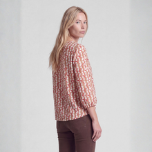 Load image into Gallery viewer, A woman with blonde hair is shown from the back wearing a beige blouse with a red and white geometric pattern, paired with brown pants, against a light gray studio background.

