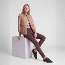 Load image into Gallery viewer, A woman models a tan quilted vest over a pink floral long-sleeve shirt, paired with dark brown skinny pants and brown suede shoes, sitting on a light gray block against a white studio background.
