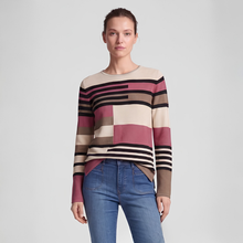 Load image into Gallery viewer, A woman modeling a long-sleeve knit sweater with horizontal and block stripes in beige, dusty rose, brown, black, and white, paired with blue jeans against a light gray studio background.
