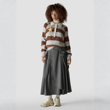 Load image into Gallery viewer, A model stands against a light gray studio background wearing a brown and cream striped knit sweater with a large gold star patch on the chest, a cream turtleneck, a long dark gray skirt, and beige sneakers.
