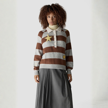 Load image into Gallery viewer, A model with curly brown hair wearing a brown and gray striped knit polo sweater with a yellow star patch, a cream turtleneck, and a dark gray pleated skirt, set against a plain light gray studio background.
