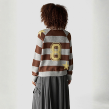 Load image into Gallery viewer, A model viewed from behind wearing a brown and gray striped knit sweater with a large gold number 8 and star motifs on the back, paired with a dark gray skirt, against a light gray studio background.
