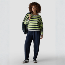 Load image into Gallery viewer, A model with curly brown hair stands against a light gray studio background wearing a green and black striped long-sleeve top, navy blue pants, and silver sneakers, with a dark jacket draped over her shoulder.
