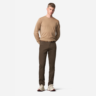 A male model stands against a light gray studio background wearing a tan long-sleeve crewneck shirt, dark brown pants, a patterned belt, and off-white sneakers.