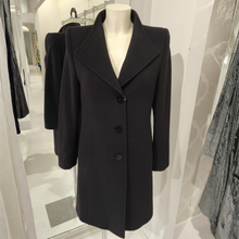 Load image into Gallery viewer, A black tailored coat displayed on a white mannequin torso in a boutique interior. The coat has a wide collar, long sleeves, and a three-button front closure.
