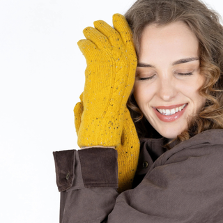 A smiling woman with closed eyes wearing a pair of mustard yellow knit gloves with small dark speckles, posed against a clean white background.