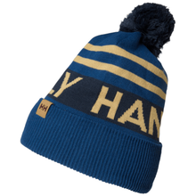 Load image into Gallery viewer, A blue and navy knit pom-pom beanie with gold stripes and 'Helly Hansen' lettering, shown on a plain white background.
