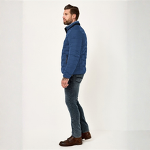 Load image into Gallery viewer, A man in profile wearing a blue quilted puffer jacket, dark blue jeans, and brown leather boots, standing against a plain white studio background.
