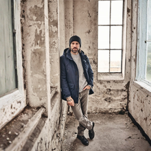 Load image into Gallery viewer, A man wearing a navy blue puffer jacket, gray sweater, beige pants, and black beanie stands in a weathered, abandoned room with peeling plaster and tall windows.
