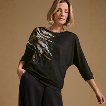 Load image into Gallery viewer, A woman wearing a black three-quarter sleeve top with a large textured, metallic silver and gold abstract pattern on the right shoulder, paired with black trousers, set against a solid warm beige background.
