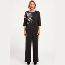 Load image into Gallery viewer, A woman modeling a black three-quarter sleeve top with a textured, metallic silver and gold abstract wing-like print on the right side, paired with black wide-leg trousers, against a light gray studio background.
