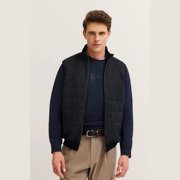 Man wearing a dark blue sweater with a vest over it on a plain background