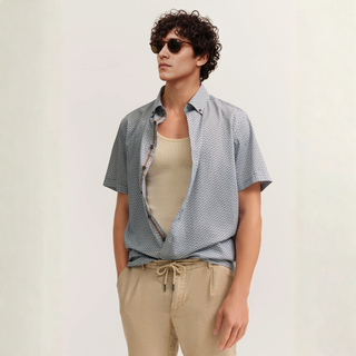 Man wearing a patterned shirt over a beige tank top with sunglasses on a plain background