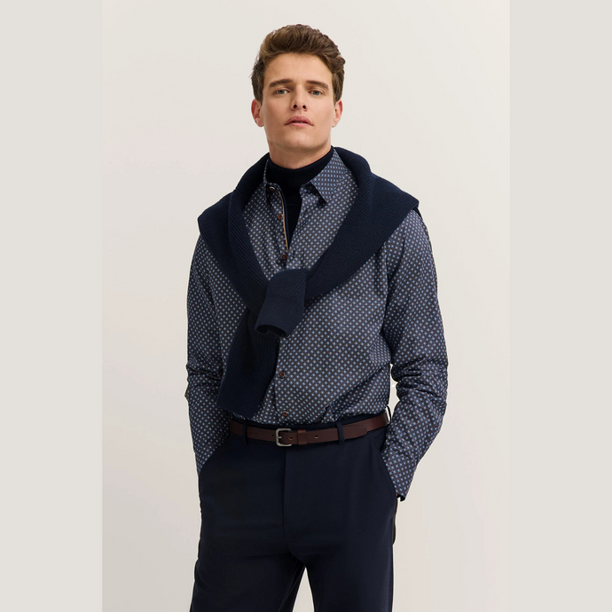 Man wearing a patterned shirt with a navy scarf and dark pants on a light background