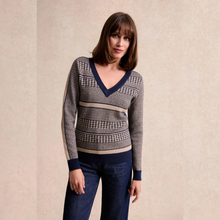 Load image into Gallery viewer, A woman modeling a V-neck knit sweater with a gray and navy houndstooth pattern and beige stripes, paired with dark blue jeans, against a textured beige background.
