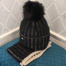 Load image into Gallery viewer, A black knit beanie hat with a large black faux-fur pom-pom and a matching striped knit scarf, displayed on a blue quilted cushion against a white paneled wall.
