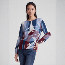 Load image into Gallery viewer, A woman modeling a long-sleeve top with an abstract geometric print in shades of blue, navy, burgundy, and white, paired with dark blue jeans against a light gray studio background.
