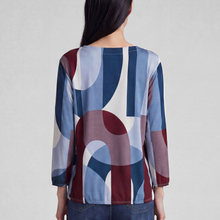 Load image into Gallery viewer, A woman viewed from the back wearing a long-sleeve blouse with an abstract geometric pattern in navy blue, burgundy, light blue, and white, paired with dark blue jeans against a light gray studio background.
