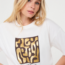 Load image into Gallery viewer, A close-up of a blonde woman wearing a white t-shirt with a large graphic print on the front. The graphic features a dark brown square containing abstract gold shapes and the text 'FLEXY' in gold.
