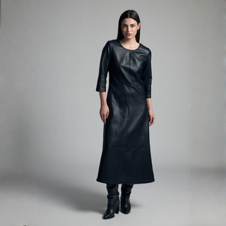 A model wearing a black leather midi dress with three-quarter sleeves and black leather boots, standing against a neutral gray studio background.