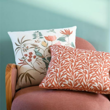 Load image into Gallery viewer, Two decorative throw pillows displayed on a dusty rose armchair against a teal wall. The pillow in the back features a botanical and bird pattern on a cream background, while the pillow in the front has a coral-orange leafy pattern on white.
